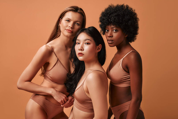 Giovani donne multietniche multirazziali in lingerie beige isolate su uno sfondo beige. Donne afroamericane, caucasiche e asiatiche in posa nello studio. Concetto di cura della pelle del viso, cosmetologia. - Foto, immagini