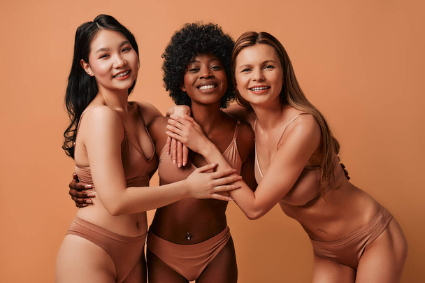 Multi-etnische multiraciale jonge vrouwen in beige lingerie geïsoleerd op een beige achtergrond. Afro-Amerikaanse, Kaukasische en Aziatische vrouwen poseren in de studio. Concept van gezichtsverzorging, kosmetologie. - Foto, afbeelding