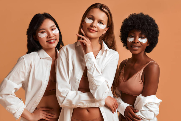 Donne multietniche in lingerie e camicie bianche isolate su sfondo beige utilizzando toppe oculari. Donne afroamericane, caucasiche e asiatiche in posa nello studio. Cosmetologia e cure termali. - Foto, immagini