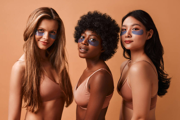 Donne multietniche in lingerie isolate su sfondo beige utilizzando toppe oculari. Donne afroamericane, caucasiche e asiatiche in posa nello studio. Cosmetologia e cure termali. - Foto, immagini