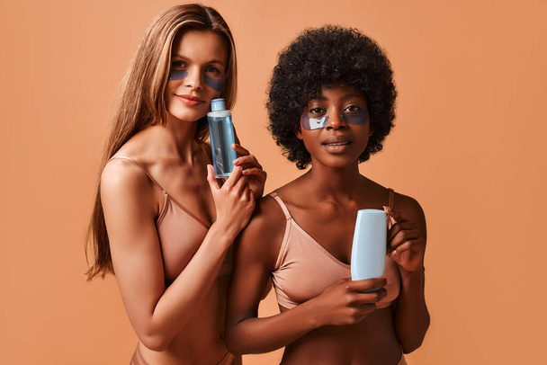 Caucasian and African-American young slim women in lingerie, using eye patches and holding bottles of beauty products, isolated on a beige background. Skin care, cosmetology and spa treatments. - 写真・画像
