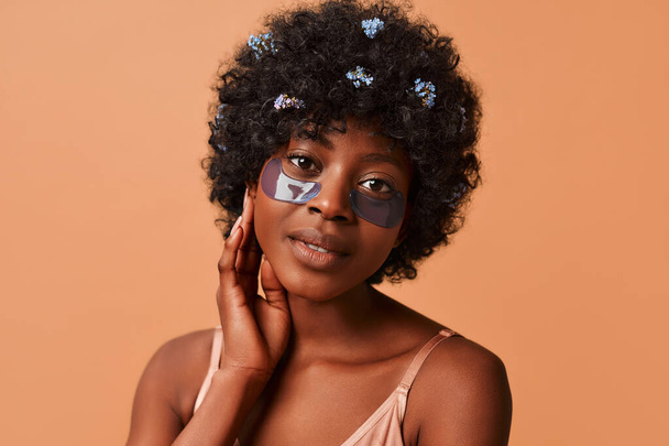 Beautiful African-American young slim woman using moisturizing eye patches in lingerie in studio on beige background. Cosmetology and spa skin care concept. - Fotografie, Obrázek