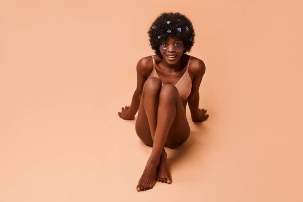 Schöne afroamerikanische junge schlanke Frau mit feuchtigkeitsspendenden Augenklappen in Dessous im Studio auf beigem Hintergrund. Kosmetologie und Wellness-Hautpflegekonzept. - Foto, Bild