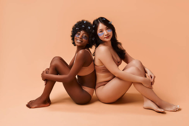 Giovani donne afro-americane e asiatiche che usano toppe sugli occhi in lingerie in posa su sfondo beige. Cosmetologia e trattamenti termali per viso e corpo. - Foto, immagini