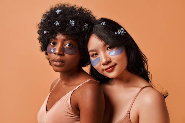African-American and Asian young women using eye patches in lingerie posing on beige background. Cosmetology and spa treatments for face and body. - Foto, Bild