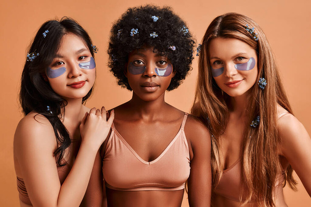 Multiethnic women in lingerie isolated on beige background using eye patches. African American, Caucasian and Asian women posing in the studio. Cosmetology and spa care. - Photo, Image