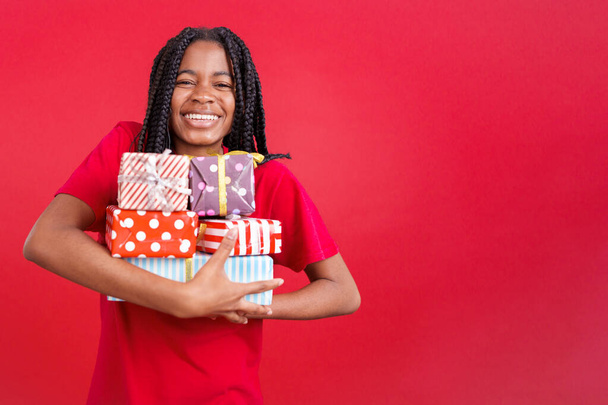 Happy african woman holding lots of colorful gifts in studio with red background - Foto, afbeelding
