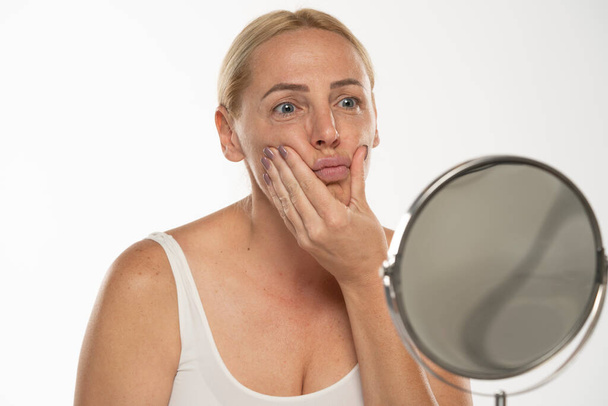 preoccupato mezza età bionda donna di fronte allo specchio tocca la bocca su sfondo bianco studio. - Foto, immagini