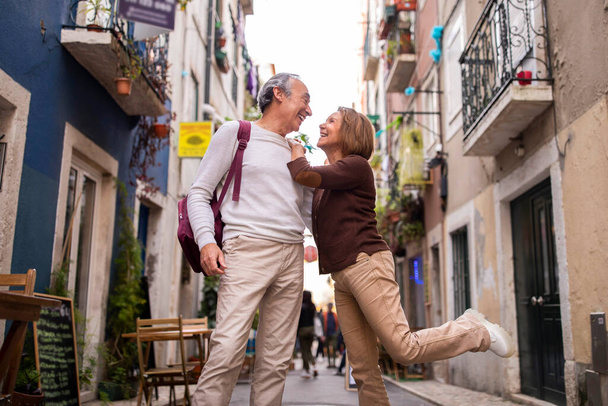休暇だ。外のリスボン通りを歩いている間に楽しいシニアカップルがポーズをとって楽しんでいます。夫の肩にもたれかかる熟女を愛し、足を持ち上げる.幸せな熟女のグロブトロッターのショット - 写真・画像