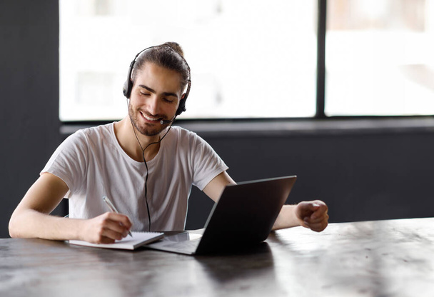 E-Learning. Jeune homme du Moyen-Orient prenant des notes apprenant à l'ordinateur portable, souriant posant avec casque assis au bureau à l'intérieur du bureau, espace libre. Homme étudiant et travaillant en ligne sur le lieu de travail - Photo, image