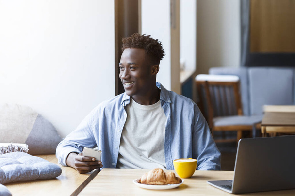 Щасливий позитивний красивий афроамериканський хлопець у повсякденному вбранні зі смартфоном та ноутбуком, насолоджуючись ранковою кавою та круасаном у кафе, дивлячись крізь вікно та посміхаючись - Фото, зображення