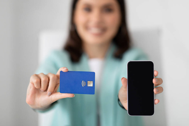 Ευτυχισμένη νεαρή Ευρωπαία γυναίκα με κοστούμι δείχνει smartphone με λευκή οθόνη, συνιστά πιστωτική κάρτα στο εσωτερικό του γραφείου, θολή, από κοντά. Επιχειρηματίας και επιχείρηση, το κέρδος με τη συσκευή και τα ψώνια - Φωτογραφία, εικόνα