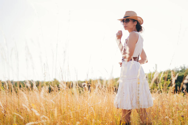 Πορτρέτο της νεαρής γυναίκας που περπατά ανάμεσα σε ψηλά χόρτα στο θερινό λιβάδι φορώντας ψάθινο καπέλο και λινό φόρεμα απολαμβάνοντας τη φύση. Αρμονία και ισορροπία. - Φωτογραφία, εικόνα