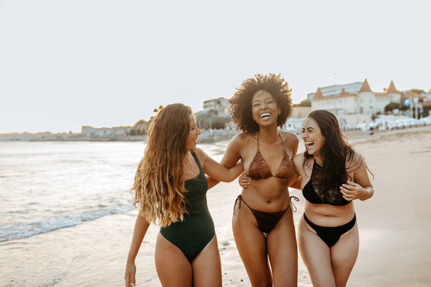 Opgewonden diverse vrouwelijke vrienden in zwemkleding omarmen en lachen vrolijk tijdens het wandelen op het strand, genieten van hun vrije tijd aan de kust van de oceaan - Foto, afbeelding