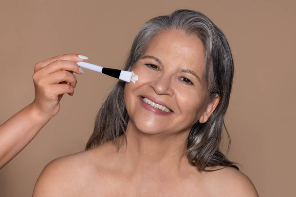Cheerful elderly caucasian lady enjoys anti-aging treatment, cosmetologist, applies cream mask with brush, isolated on brown background. Beauty care, wrinkle in menopause, moisturizing cosmetics - Photo, Image