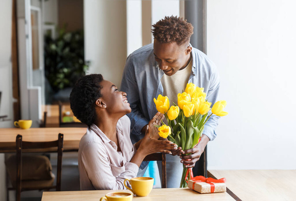 Piękne szczęśliwe młode, wesołe czarne pary mają romantyczną randkę w restauracji. Kochający Afrykanin amerykański facet przynosząc kwiaty i pudełko prezentów do jego dość podekscytowany dziewczyna kawiarnia wnętrze - Zdjęcie, obraz