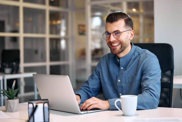Jonge bebaarde knappe zakenman in bril en shirt zittend in een modern kantoor met een laptop met een kopje koffie. Succesvol hipster team in coworking. Freelancers. - Foto, afbeelding