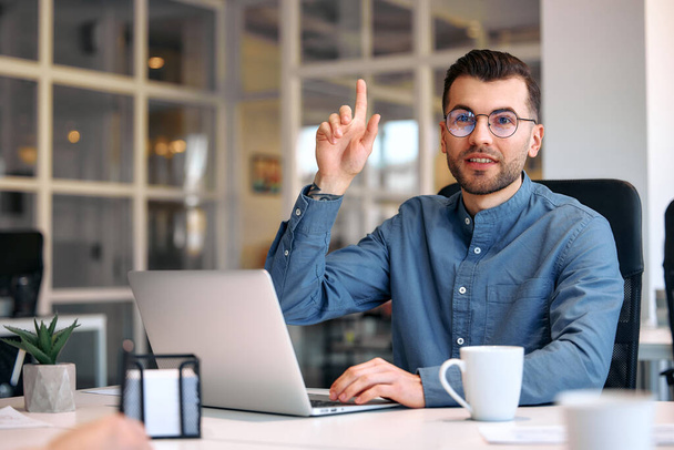 Νεαρός γενειοφόρος όμορφος επιχειρηματίας με γυαλιά και πουκάμισο κάθεται σε ένα σύγχρονο γραφείο με ένα φορητό υπολογιστή με ένα φλιτζάνι καφέ. Επιτυχημένη ομάδα χίπστερ στη συνεργασία. Ελεύθεροι επαγγελματίες. - Φωτογραφία, εικόνα