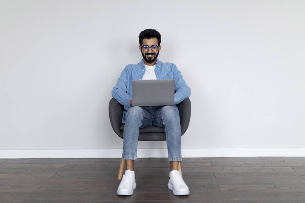 Sikeres indiai férfi Szabadúszó Laptop dolgozik Online, miközben ül a szék felett szürke háttér otthon, jóképű keleti fickó számítógép használata, Böngészés Internet a kommunikáció, Másolás tér - Fotó, kép