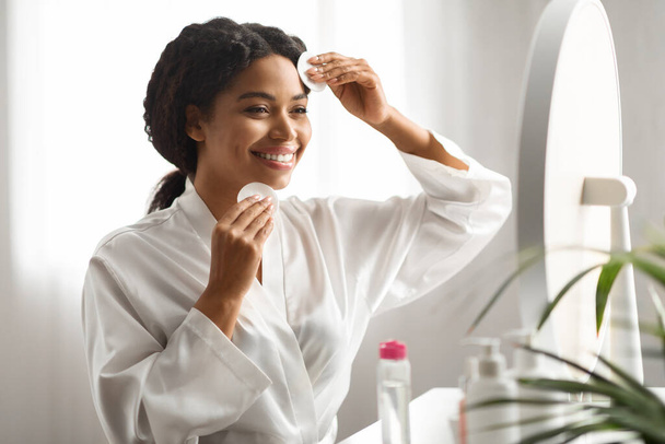 Mujer sonriente negra usando almohadillas de algodón, Cara de limpieza con agua micelar en casa, Atractiva mujer afroamericana cuidando la piel, Disfrutando de la rutina diaria de cuidado facial de la piel, Primer plano - Foto, imagen