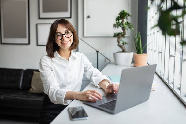Successful freelancer. Happy young lady sitting at desk with laptop, cheerful european woman smiling and posing at camera while working on computer in cozy office, free space - 写真・画像