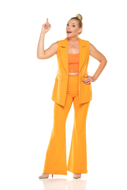 Middle aged smiling senior woman in yellow sleevless jacket and trousers pointing up on a white studio background - Foto, Imagem