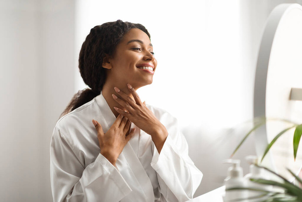 Neck Lift Concept. Smiling African American Woman In Silk Robe Touching Her Soft Smooth Skin While Looking In Mirror At Home, Happy African American Female Enjoying Result Of Beauty Treatments - Photo, Image