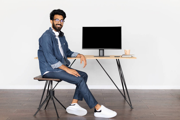 Positivo sonriente joven guapo hombre de Oriente Medio en traje casual sentado en el escritorio con ordenador PC con pantalla vacía negro. Árabe chico trabajando en línea desde el hogar oficina, maqueta, espacio de copia - Foto, imagen