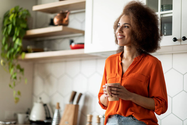 Jó reggelt! Portré mosolygó afro-amerikai nő ivás kávé a konyhában, vidám fekete nő áll egy csésze forró ital a kezében, és néz félre, élvezi a hazai pihenés - Fotó, kép