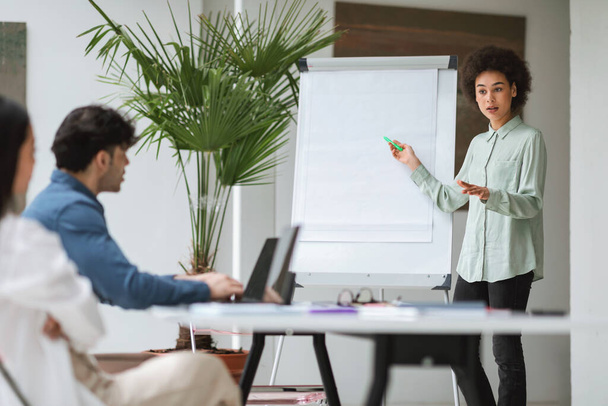 Business Presentation. Black Businesswoman Presenting Strategy Ideas To Young Engaged Colleagues Of Diverse Ethnicities At Corporate Meeting In Office Interior, Pointing At Whiteboard During Speech - Foto, imagen