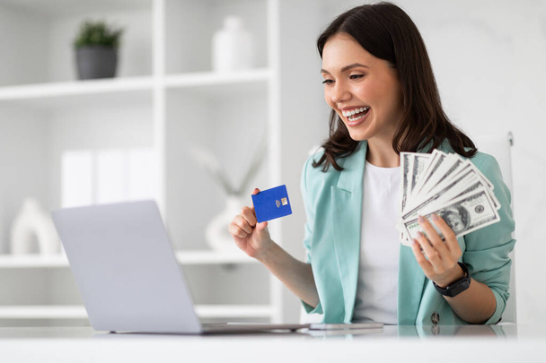 Happy excited young european lady in suit look at laptop, show credit card, many money cash in office interior. Businesswoman and successful business, work, win with device and profit for startup - Photo, Image
