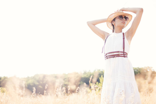 Geest van vrijheid. Een aantrekkelijk boho meisje in bloes, hoed en zonnebril op het veld op de achtergrond van een blauwe lucht. Zomervakantie, reizen. Boheemse, moderne hippie stijl - Foto, afbeelding
