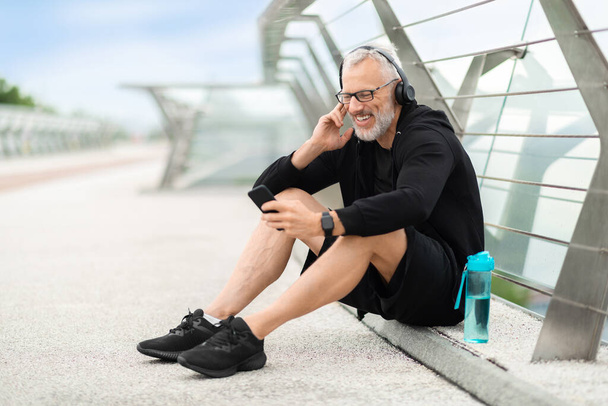 Sportif âgé assis sur le pont, boire de l'eau, faire une pause en faisant du jogging en plein air. Homme sportif senior utilisant un smartphone, appuyant sur le bouton sur les écouteurs sans fil, écoutant de la musique - Photo, image