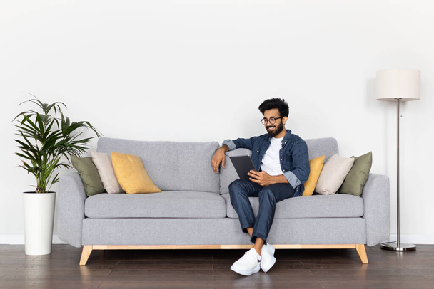 Bonito cara oriental usando tablet digital enquanto relaxa no sofá em casa. Millennial árabe homem navegando novo aplicativo, trabalhando on-line ou fazer compras na Internet, sentado na sala de estar, espaço livre - Foto, Imagem