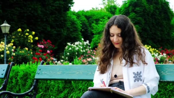 Egy fiatal nő tollal ír egy füzetben, miközben egy padon ül a parkban.. - Felvétel, videó