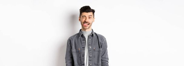 Homme drôle avec moustache montrant la langue et cligner des yeux, vous faisant rire, debout sur fond blanc. - Photo, image