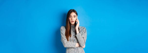 Shocked girl talking on mobile phone, gasping amazed at camera, speaking with someone and hear shocking news, standing in dress on blue background. - Фото, зображення