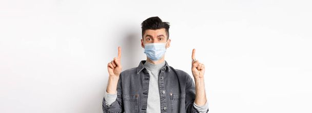 Pandemian elämäntapa, terveydenhuolto ja lääketiede. Tyylikäs moderni mies lääketieteellisessä naamiossa, joka näyttää mainosta, osoittaa sormella logoa, seisoo valkoisella taustalla. - Valokuva, kuva