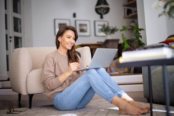 Portrait of beautiful long haired woman wearing casual clothes while using laptop for work at home. Confident female sitting on the floor and having video call. - Фото, зображення
