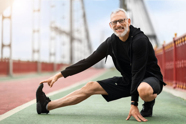 Alegre anciano sano estirando las piernas y sonriendo en el puente de la ciudad. Deportista senior, corredor al aire libre, motivación para el fitness, energía y entrenamiento activo en la calle, espacio para copiar - Foto, imagen