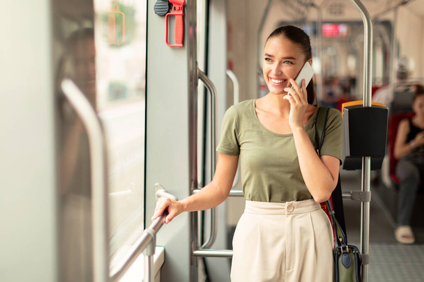 Ευτυχισμένη κυρία των επιβατών μιλώντας στο κινητό στέκεται στο σύγχρονο τραμ μέσα, κοιτάζοντας μακριά. Νεαρή γυναίκα απολαμβάνει βόλτα στα μέσα μαζικής μεταφοράς, επικοινωνώντας ενώ κινείται προς την εργασία. Αντιγραφή χώρου - Φωτογραφία, εικόνα