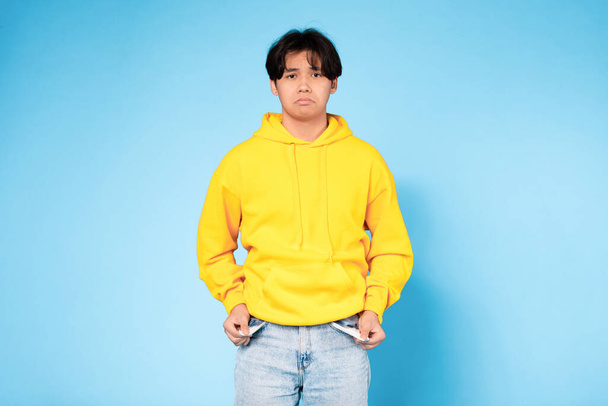 Poverty Concept. Upset Asian Young Teen Guy Showing His Empty Pockets And Looking At Camera, Having Financial Issues Standing Over Blue Studio Background. No Money, Crisis - Фото, зображення