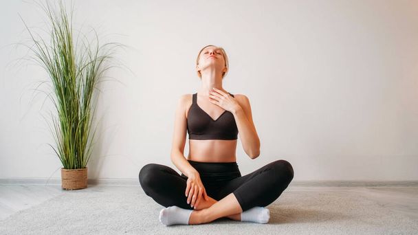 Morning exercise. Home fitness. Indoor training. Relaxed inspired athletic girl stretching neck muscles sitting cross-legged on floor at light studio interior copy space background. - Foto, imagen