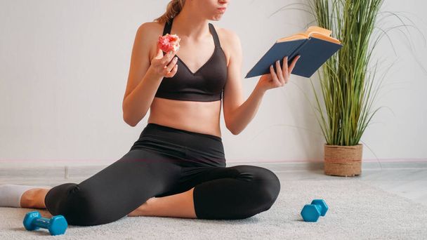 Diet study. Fitness nutrition. Junk food calorie. Unrecognizable athletic woman reading book eating sweet doughnut dessert on floor with dumbbells on light free space. - Foto, Imagem