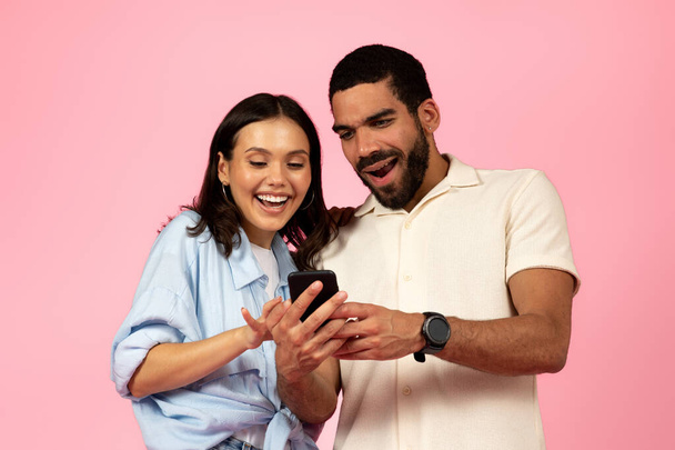 幸せな陽気な愛する若い多人種のカップルのヒスパニック系の男とブルネットの女性を使用してスマートフォン上でピンク色のスタジオの背景,チェックしてください素晴らしいモバイルアプリ,オンラインアシスタント,チャットボット - 写真・画像