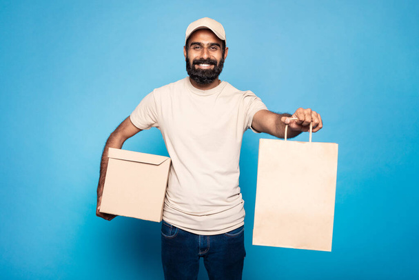 Verkkokauppa ja jakelupalvelu. Positiivinen intialainen kuriiri, joka antaa ostoskassin ja hymyilee kameralle, seisoo sinisellä studion taustalla, vapaa tila - Valokuva, kuva