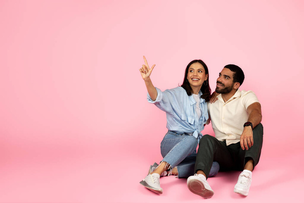 Glücklich liebendes junges hispanisches Paar, das auf dem Boden sitzt und sich umarmt, auf den Kopierraum für Werbung oder Text zeigend, isoliert auf buntem rosa Studiohintergrund. Tolles Reiseangebot für zwei - Foto, Bild
