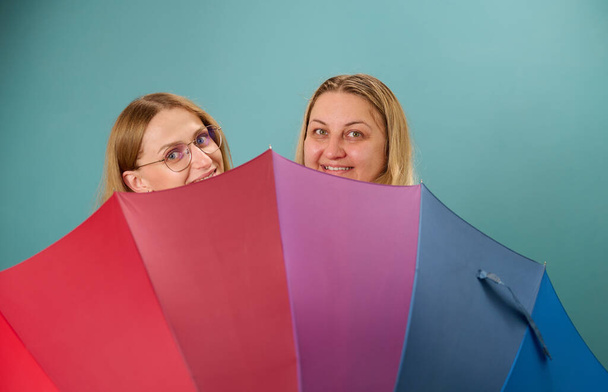 Дві щасливі молоді усміхнені лесбіянки обіймаються за барвистою парасолькою. Приваблива небайдужа жіноча підтримка, лесбійська пара стоїть і обіймає. - Фото, зображення