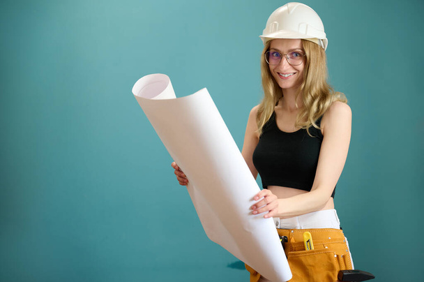 Young architect girl with protective helmet holding a house plan in hands isolated on background. Pretty caucasian female architect are planning new project. People work on architecture project - Photo, Image
