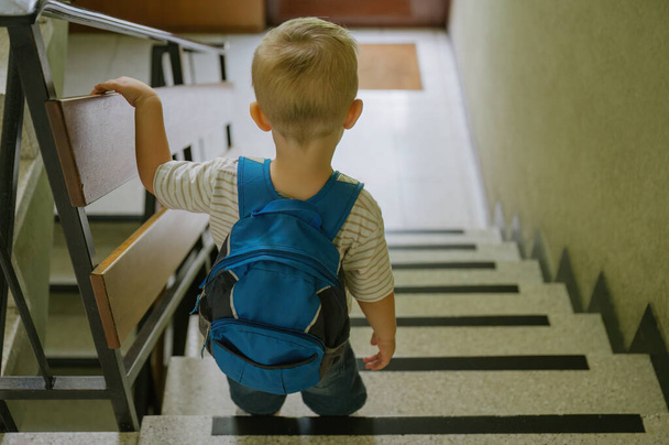 Widok z tyłu małego chłopca z blond włosami w luźnych ubraniach z plecakiem stojącym na schodach przed pójściem rano do przedszkola. koncepcja rodziny - Zdjęcie, obraz